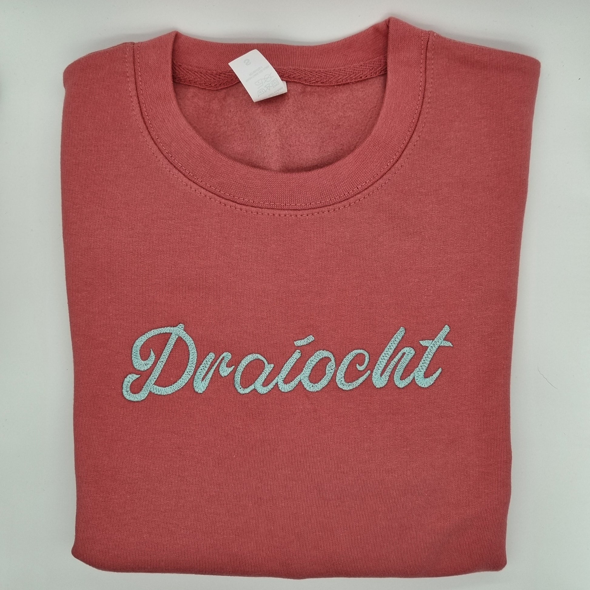 Red/pink folded sweatshirt with "Draíocht" embroidered onto it with light blue thread